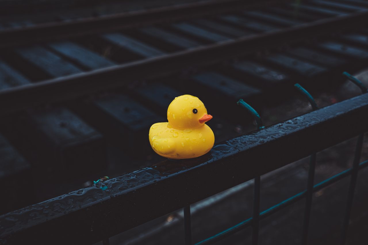 A vibrant yellow rubber duck sits on a wet black railing near railway tracks, conveying whimsy.